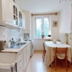 Biela retro kuchyňa s antikorovým drezom a jedálenským stolom s obrusom.