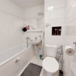 Pôvodná kúpeľňa s bielym obkladom, klasikou vaňou, umývadlom a kombinovaným WC s čiernym koberčekom.