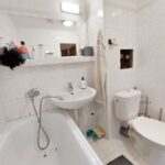 Staršia kúpeľňa obložená bielymi kachličkami s vaňou, zrkadlom a policou s hygienickými potrebami.