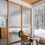 Moderný interiér s drevenou deliacou priečkou, žltým čalúneným kreslom a veľkoformátovým oknom do zasneženého lesa.