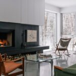 Moderný čierny kozub, sklenený konferenčný stolík a drevené hojdacie kreslo pri okne.