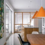 Kuchynský ostrov s dreveným obkladom, oranžové závesné lampy a jedálenský stôl s dizajnovými stoličkami.