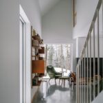 Interiér s bielymi stenami, vysokým otvoreným stropom a štýlovým zeleným kreslom pri schodisku.