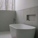 Vaňa v minimalistickej kúpeľni so svetlosivým mozaikovým obkladom a oknom s výhľadom na les.