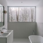 Kúpeľňa v bielej a svetlosivej farbe s vaňou, dvojumývadlom a širokým oknom s výhľadom na zasnežený les.