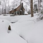 Rodinný dom so sedlovou strechou, drevenou fasádou a veľkoformátovými oknami na kopci s potokom.