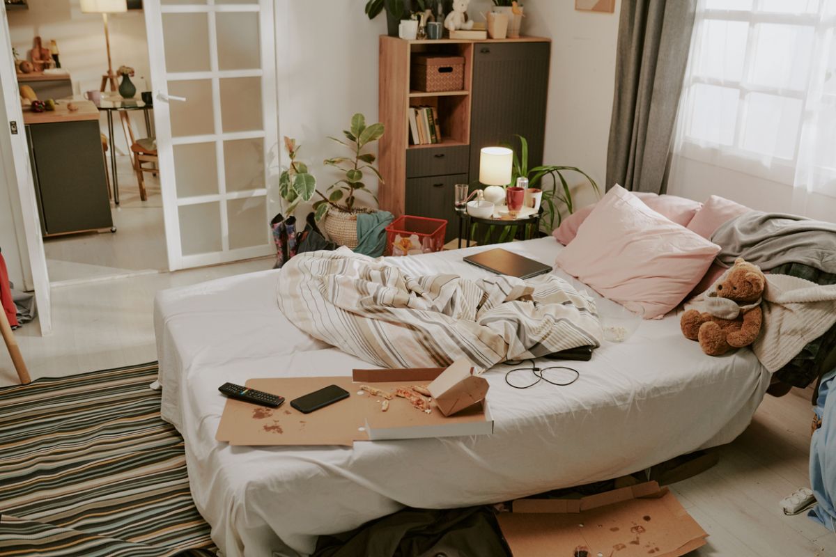 Neupravená posteľ v spálni s krabicou od pizze, zvyškami jedla a rozhádzaným oblečením.