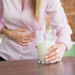 Žena v ružovej košeli drží v jednej ruke pohár mlieka a druhú má položenú na bolestivom bruchu.