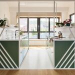 Symetrická kuchynská linka so zelenými skrinkami, bielou pracovnou doskou a bielym oceľovým rámom v loftovom interiéri.