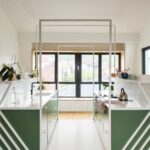 Symetrická kuchyňa so zelenými skrinkami, bielou pracovnou doskou a bielym kovovým rámom pri okne.