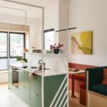 Zelená kuchynská skrinka so zelenými čelami a bielou pracovnou doskou, za ktorou je jedálenský kút s červenou lavičkou a dreveným stolom.