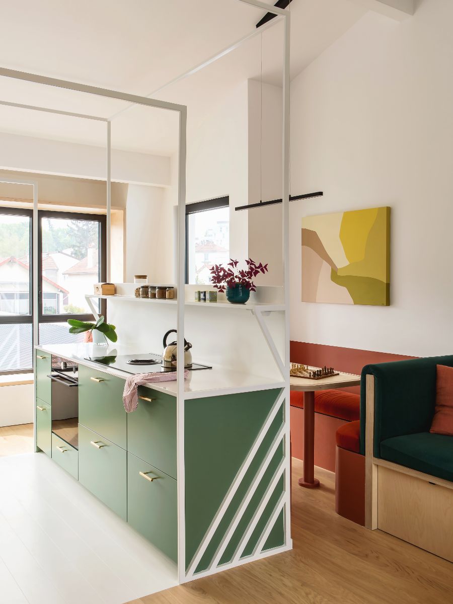 Zelená kuchynská skrinka so zelenými čelami a bielou pracovnou doskou, za ktorou je jedálenský kút s červenou lavičkou a dreveným stolom.