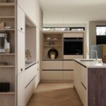 Kuchynský kút s vysokými béžovými skriňami, vstavanou čiernou rúrou a dubovými parketami v tvare rybej kosti.