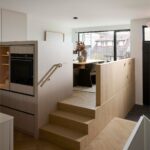 Svetlé drevené schodisko prepojené s minimalistickým kuchynským ostrovom a vyvýšenou jedálenskou zónou.