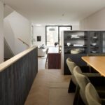 Priehľad z jedálne cez drevenú chodbu do svetlej obývacej izby s tmavosivou knižnicou a bielym nábytkom.