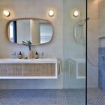 Ratanová konzolová skrinka pod oválnym dreveným zrkadlom v kontraste s modrým obkladom sprchovacieho kúta.