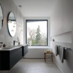 Čierne umývadlové skrinky pod kruhovými zrkadlami v kúpeľni s voľne stojacou vaňou pred panoramatickým oknom.