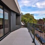 Priestranný strešný balkón s hladkou svetlou podlahou, čiernym kovovým zábradlím a tmavým dreveným obkladom steny.