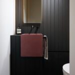 Matné vínové umývadlo na stene s vertikálnym čiernym obkladom s minimalistickou čiernou batériou a úzkym zrkadlom.