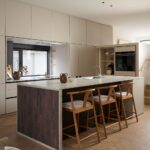 Moderný kuchynský ostrov s krémovou pracovnou doskou, tmavým dreveným čelom a tromi dubovými barovými stoličkami.