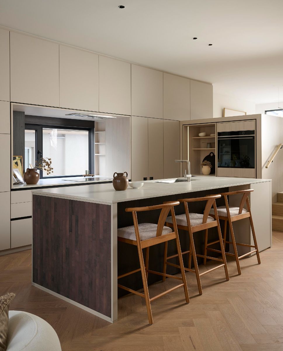 Moderný kuchynský ostrov s krémovou pracovnou doskou, tmavým dreveným čelom a tromi dubovými barovými stoličkami.