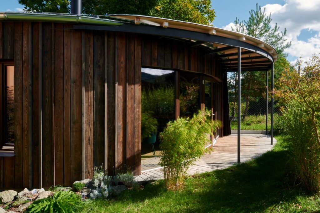 Exteriér moderného drevodomu s oblými líniami a drevenou terasou.
