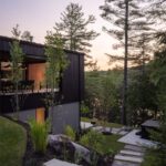 Moderný dom s čiernym dreveným obkladom, betónovým suterénom, kamenným chodníkom a exteriérovým osvetlením.