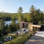 Moderný čierny dom s dreveným obkladom, priestrannou terasou, kamenným ohniskom a okolitým ihličnatým lesom.