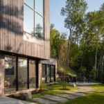 Poschodový dom s veľkými oknami, kamenným chodníkom, trávnatou záhradou a lesným pozadím.