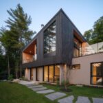 Moderný dom s drevenou fasádou, osvetleným interiérom, kamennou cestičkou v trávniku a vysokými ihličnanmi.