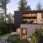 Časť moderného domu s drevenou fasádou, osvetleným interiérom, chodníkom v tráve a posedením.