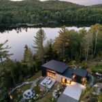 Letecký pohľad na čierny dom s plochou strechou, kamenným chodníkom a blízkym jazerom uprostred lesa.