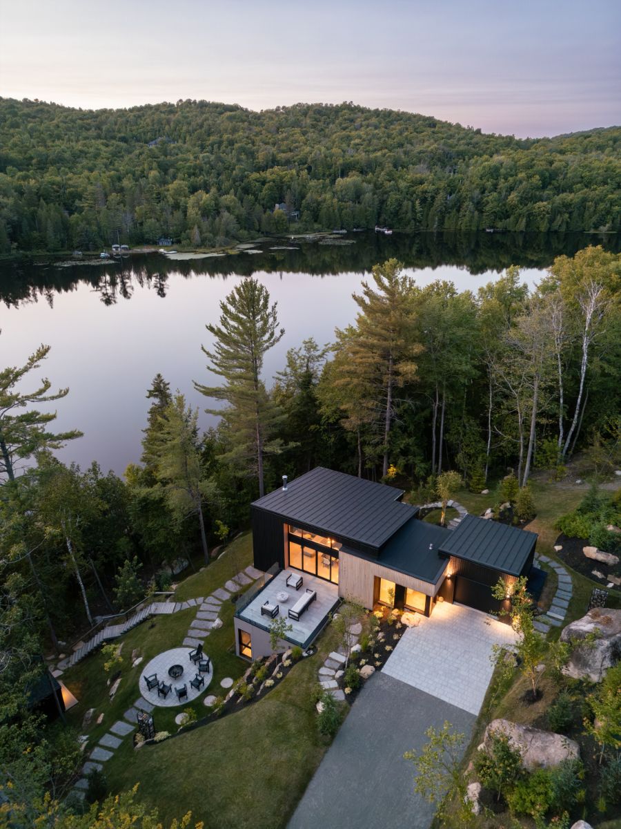 Letecký pohľad na čierny dom s plochou strechou, kamenným chodníkom a blízkym jazerom uprostred lesa.