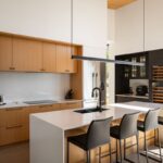 Moderný kuchynský ostrov s bielou doskou, čiernou batériou, drevenými skrinkami a tmavou vinotékou v pozadí.