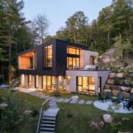 Osvetlená moderná vila v svahu s kamenným ohniskom, dreveným schodiskom a hustým zmiešaným lesom.