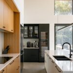 Čierna vstavaná vinotéka s kávovarom, biely kuchynský ostrov a veľké okno s výhľadom na zelené stromy.