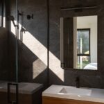 Kúpeľňa s tmavým kamenným obkladom, drevenou skrinkou, bielym umývadlom a čiernou sprchovou hlavicou.