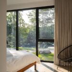 Minimalistická spálňa s drevenou posteľou, svetlým závesom, čiernym kreslom a výhľadom do lesnej záhrady.