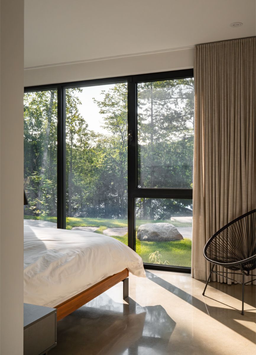 Minimalistická spálňa s drevenou posteľou, svetlým závesom, čiernym kreslom a výhľadom do lesnej záhrady.