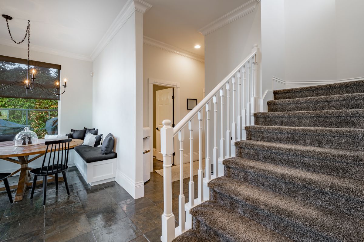 Drevený okrúhly stôl, čierna latková stolička, biela rohová lavica a schodisko s hnedým kobercom.