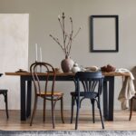 Masívny drevený stôl s čiernou kovovou podnožou, tmavé ohýbané stoličky a béžová stena s čiernym rámom.