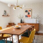 Moderný drevený stôl, žlté čalúnené stoličky, geometrická tapeta a mosadzný luster s bielymi guľami.
