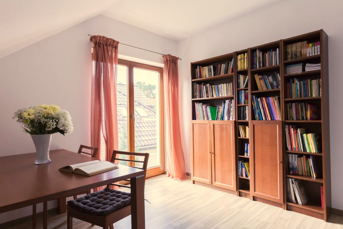 Tmavohnedý jedálenský stôl, vysoká drevená knižnica, svetlá podlaha a marhuľové závesy na balkónovom okne.