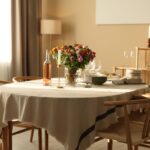 Okrúhly stôl s béžovým obrusom, svetlé drevené stoličky s výpletom a váza s pestrými kvetmi.