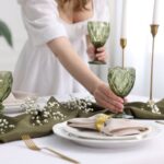 Biely prestretý stôl, zelené brúsené poháre, zlatý príbor a olivovo zelený textilný behúň.