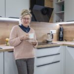 Žena v bielej kuchyni s dubovou pracovnou doskou, čiernou varnou doskou a moderným sodastreamom v rohu linky.