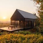 Malá moderná čierna chata s veľkou drevenou terasou a skleneným zábradlím pri západe slnka v lese.