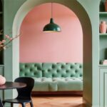 Pohľad cez oblúkový priechod v pastelovozelene knižnici na zelenú sedačku pri ružovej stene.