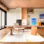 Svetlý moderný interiér s drevenou podlahou, nízkym kreslom, geometrickým kobercom a betónovým stropom v pastelovom odtieni.