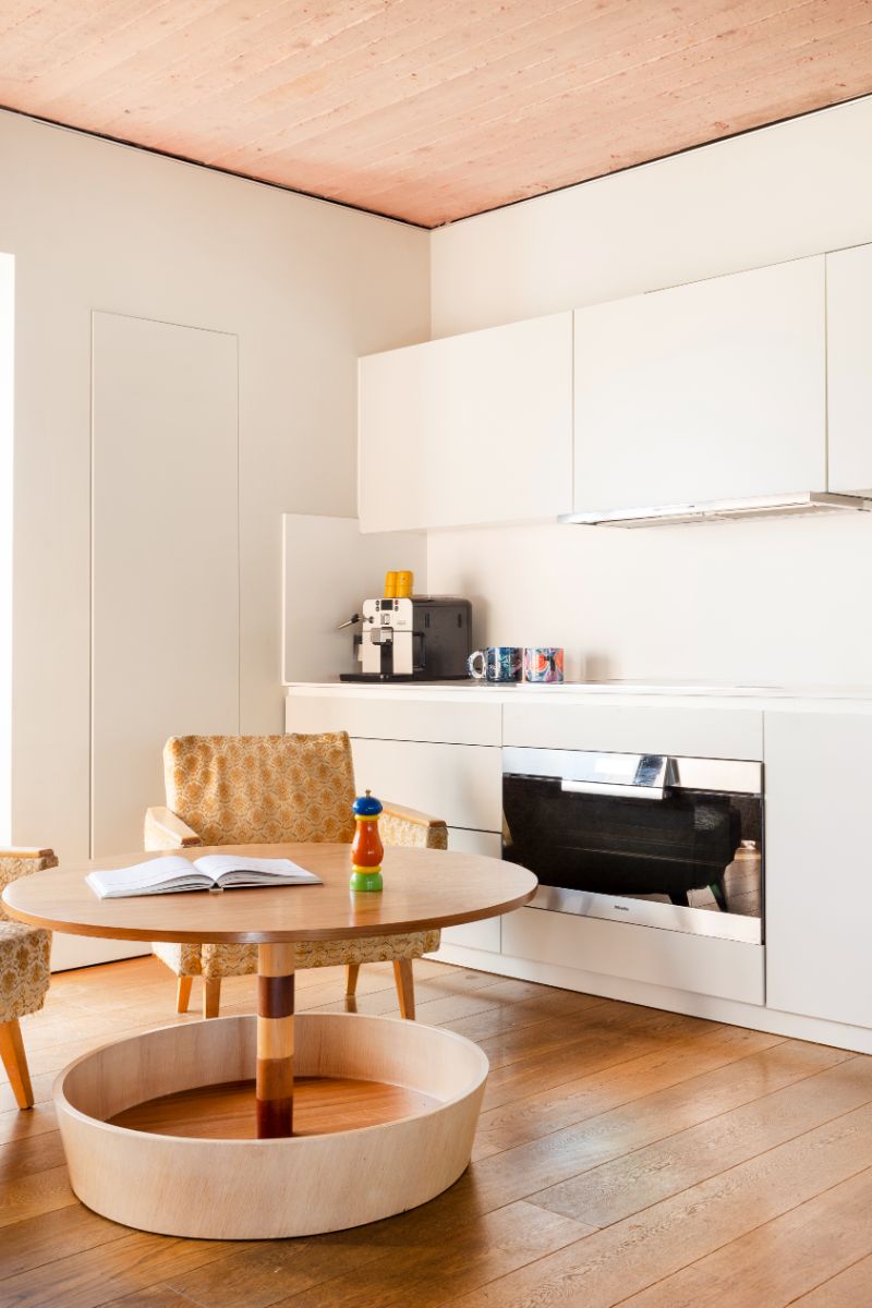 Kuchynský kút s bielymi skrinkami, okrúhlym dreveným stolíkom a vzorovaným kreslom v retro štýle.